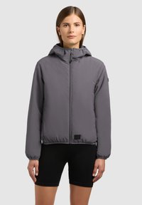 Mlada ženska, oblečena v sivo kapucar z zadrgo in elastičnimi manšetami ter črne kolesarske hlače, stoji pred enobarvnim svetlim ozadjem.