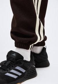 Μαύρα παπούτσια Adidas με τρεις ανακλαστικές ρίγες, φορεμένα με λευκές κάλτσες και καφέ φόρμες με λευκές πλευρικές ρίγες.