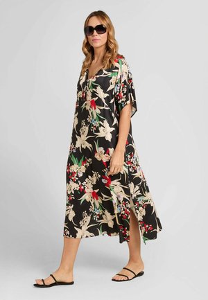 Donna che indossa un abito midi nero con stampa floreale e spacco laterale, sandali neri e grandi occhiali da sole scuri su sfondo chiaro e uniforme.