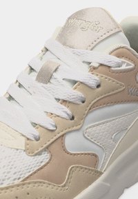 Κοντινό πλάνο μπεζ και λευκού αθλητικού παπουτσιού Wrangler με υφή στο πλαϊνό πάνελ, λευκά κορδόνια και ανάγλυφο λογότυπο στη επενδυμένη γλώσσα.