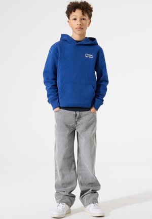 Sudadera de polar azul con bolsillo frontal y letras blancas; combinada con jeans sueltos de color gris claro y zapatillas blancas. Fondo blanco liso.