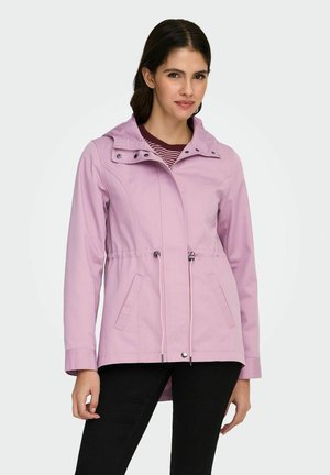 Femme portant une veste à capuche violette claire avec cordons de serrage, poches avant, et pantalon noir, debout devant un fond uni.