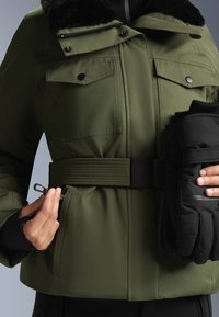 Olivno zeleno jakno s krznom obloženim ovratnikom, sprednjimi žepi in zategnjenim pasom. Črne tople rokavice v roki.