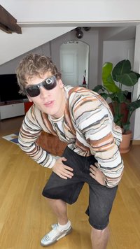 Mudel mitme triibuga soe, mustades lühikestes pükstes ja hõbedastes spordijalanõudes. Kandmas päikeseprille, poseerib käed puusas moodsas sisehaldas.