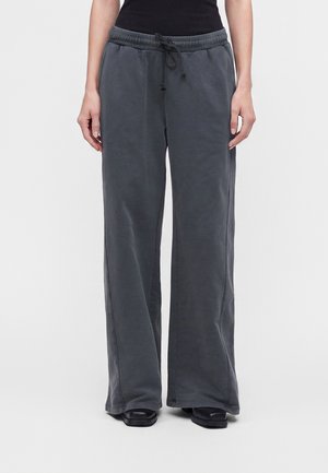 Personne portant un pantalon de survêtement ample gris foncé avec taille à cordon et chaussures noires, debout devant un fond clair uni.