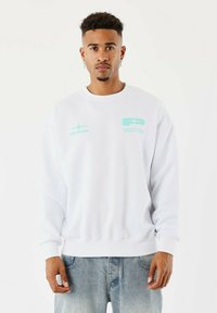 Hombre con cabello corto y rizado, vistiendo un sudadera blanca con un gráfico en azul claro y jeans azul claro, de pie contra un fondo blanco.