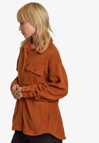 Burnt orange fleece shirt jacket with a button closure and chest pocket. Features rounded hem and relaxed fit with dropped shoulders.