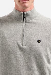 Grauer Reißverschluss-Pullover mit hohem Kragen, geripptem Hals und einem sechseckigen Logo-Patch auf der Brust, hergestellt aus einem weichen Textilgewebe.