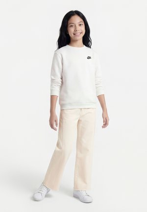 Glimlachend kind met lang zwart haar, een witte Nike-trui, beige broek en witte sneakers, staand op één voet tegen een effen achtergrond.