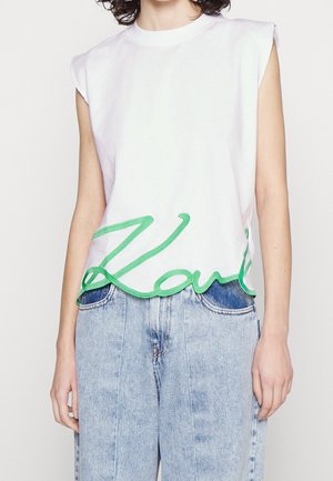 Biały bawełniany top bez rękawów z zielonym haftowanym napisem przy dole. Stylizowany z jasnoniebieskimi jeansami.