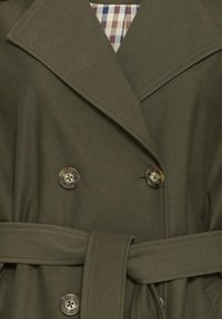 Manteau croisé vert olive foncé avec larges revers, quatre boutons, ceinture assortie et doublure intérieure à carreaux visible au niveau du col.