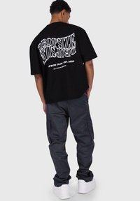 Zwarte oversized t-shirt met witte grafische tekst op de rug, gecombineerd met losse grijze cargobroek en witte sneakers.