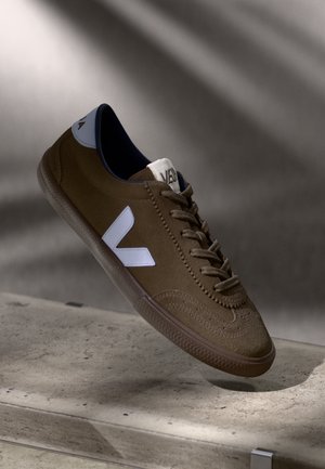 Sneaker in suede marrone con logo bianco, punta rotonda e suola in gomma testurizzata; presenta cuciture decorative e dettagli azzurri sul tallone.