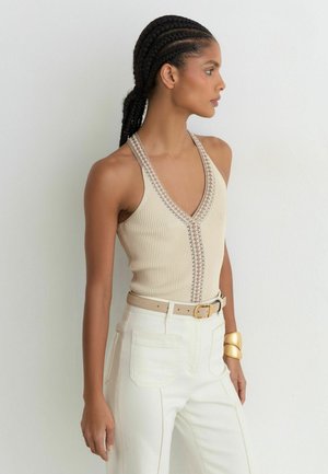 Femme aux cheveux tressés portant un top halter côtelé beige, un pantalon blanc taille haute, une ceinture beige et un bracelet doré, debout de profil.