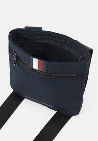 Borsa a tracolla navy con zip superiore e tasca frontale. Presenta un logo in testo bianco e un accento rigato in rosso, bianco e blu. Texture liscia.