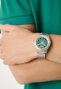 Ceas din oțel inoxidabil argintiu cu un cadran geometic turcoaz, accente aurii și o brățară legată. Purtați pe un încheietură cu țesătură verde.