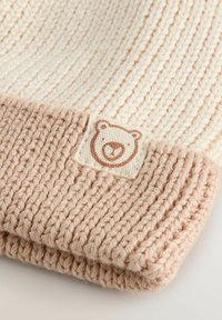 Strikket beige og lysebrun hue med ribbestrikket kant og syet på patch med en simpel bjørneansigt illustration.