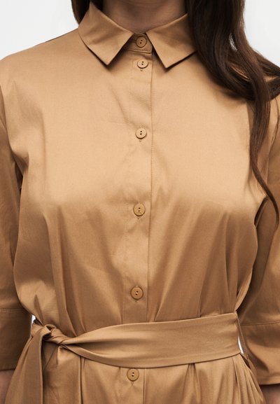 Femme portant une robe boutonnée marron clair avec un col et une ceinture en tissu assortie nouée à la taille, avec de longs cheveux bruns visibles.