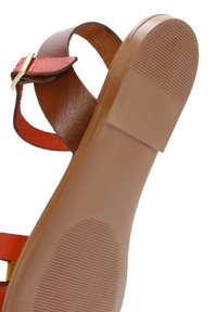Sandalia de cuero marrón con tira roja y hebilla dorada. Suela plana de goma con patrones de agarre texturizados. Diseño abierto para mayor transpirabilidad.