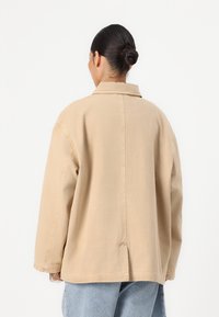 Beige oversized jacket with a classic collar, long sleeves, and side slits at the hem, crafted from a soft, durable cotton blend.