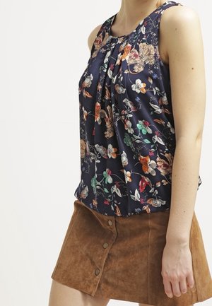 Donna che indossa un blusa senza maniche con fantasia floreale blu navy e una gonna marrone in suede con bottoni, in piedi di fronte a uno sfondo bianco semplice.