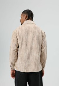 Beige corduroy overhemd met verticale strepen, lange mouwen, klassieke kraag en afgeronde zoom, gezien van achteren met een zwarte tailleband.