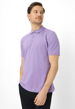 Mann trägt ein hellviolettes Poloshirt mit kurzen Ärmeln und schwarze Hose, steht mit den Händen in den Taschen und schaut seitlich vor weißem Hintergrund.