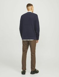 Marinblå stickad tröja med rund halsringning, bärs över en vit skjorta, kombinerad med bruna byxor och svarta skor, sedd bakifrån.