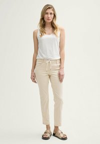 Top con scollatura a fentaglio bianco abbinato a pantaloni beige con coulisse e sandali beige con accenti neri, in piedi contro uno sfondo neutro.