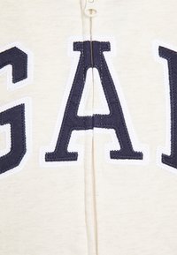 Felpa con zip color crema caratterizzata da grandi lettere ricamate in blu navy "GAP" con bordi bianchi, texture liscia e una zip centrale.