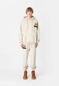 Sudadera beige con un parche de oso de peluche, bolsillo frontal y acentos grises, combinada con pantalones de chándal beige a juego y botas marrones con cordones.