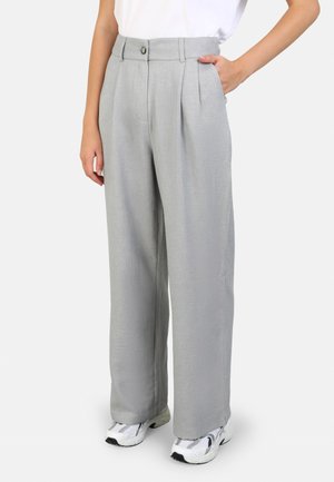 Osoba ubrana w jasnoszare spodnie o szerokich nogawkach z przednimi plisami, zapięciem na guzik w pasie, białe trampki i białą koszulę, stojąca z jedną ręką w kieszeni.