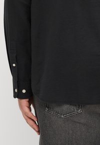 Schwarzes Langarmshirt mit abgerundetem Saum, knöpfbaren Manschetten mit perlenähnlichen Knöpfen und einer glatten Baumwolltextur. Kombiniert mit grauen Jeans.