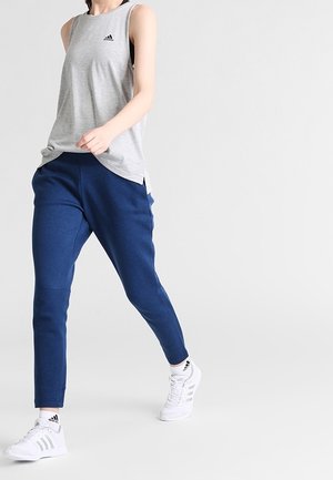 Person, der spaziert, trägt ein graues ärmelloses Adidas-Oberteil, blaue Jogginghose und weiße Adidas-Sneaker mit Logo auf den Socken.