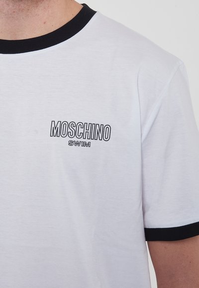T-shirt bianco in cotone con colletto e rifiniture delle maniche a costine nere. Presenta la scritta "MOSCHINO SWIM" in nero sulla parte sinistra del petto.