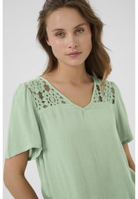 Blouse vert menthe à manches courtes, décolleté en V et tissu texturé. Comprend un motif floral en dentelle sur les épaules. Design doux et léger.
