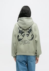 Hosszú sötét hajú személy sage zöld kapucnis felsőt visel, fekete pillangóval és "OBEY" felirattal a hátán, kék farmernadrággal párosítva, háttal áll.