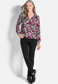 Bunt gemusterte Bluse mit geometrischem Muster in Schwarz, Pink und Hellblau, kombiniert mit schwarzen Slim-Fit-Hosen und schwarzen Sneakers.