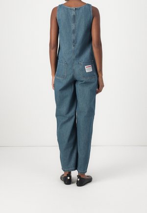 Osoba stojąca, ubrana w bezrękawnikowy niebieski kombinezon z denimu z zamkiem z tyłu, dwoma tylnymi kieszeniami oraz czarnymi butami na białym tle.