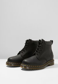 Botas de tobillo de cuero negro con punteras redondas, frente con cordones y costuras amarillas a lo largo de las suelas. Acabado suave y suelas de goma resistentes.