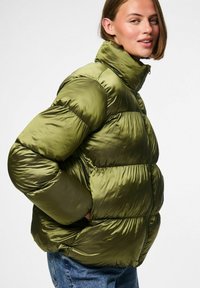 Žalias pūkuotas džemperis su aukštu apykakle, turintis blizgią, siuvinėtą tekstūrą ir kvadratą formą. Patogus dizainas su šoninėmis kišenėmis patogumui.