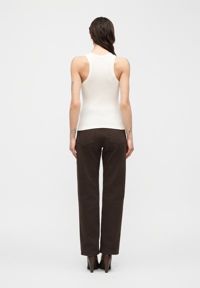 Λεπτός άνθρωπος που φοράει λευκή αμάνικη μπλούζα, σκούρο καφέ παντελόνι με ίσια γραμμή και ψηλοτάκουνα παπούτσια, εμφανίζεται από πίσω με φόντο ένα απλό φόντο.