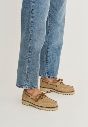 Voeten in beige suède bootschoenen met bruine veters en lichtblauwe rechte jeans tegen een effen achtergrond.