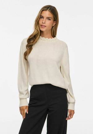 Femme aux longs cheveux portant un pull en maille crème avec un col festonné et un pantalon noir taille haute, debout devant un fond uni.