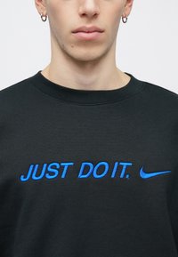 Μαύρο φούτερ με μπλε κεντημένο κείμενο «JUST DO IT.» στην πρόσοψη και λογότυπο Nike swoosh. Ομαλή υφή και κλασικός σχεδιασμός με λαιμόκοψη crew.