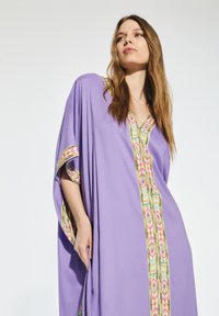 Lila kaftan med mönstrad kant i rosa, grön och gul. Löst sittande, V-ringad design och vida ärmar. Mjuk tygtextur.