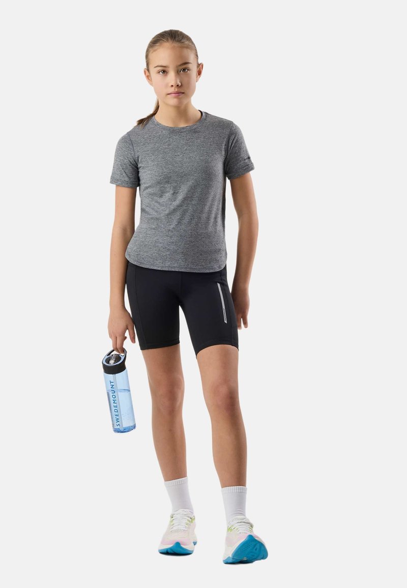 Fille portant un t-shirt gris, un short noir, des chaussettes blanches et des baskets, tenant une bouteille d'eau bleue sur un fond uni.