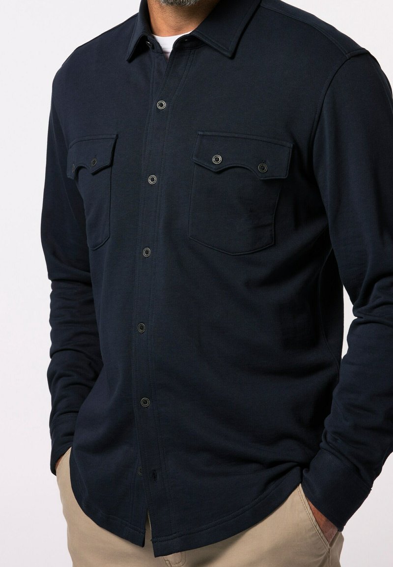Homme portant une chemise bleue marine boutonnée avec deux poches poitrine et un pantalon beige, les mains dans les poches, sur un fond uni.