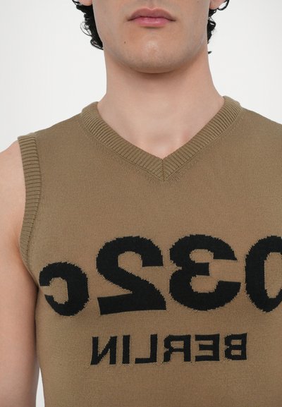 Brauner gestrickter ärmelloser Pullover mit V-Ausschnitt, auf dessen Vorderseite in fetter schwarzer Schrift "032C BERLIN" deutlich zu sehen ist. Glatte Textur.