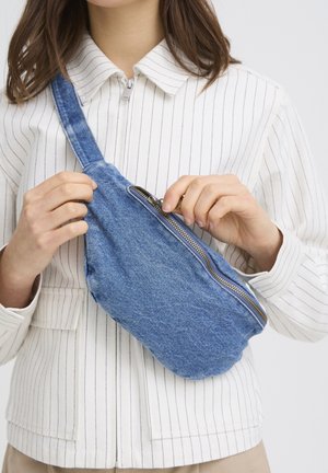 Fehér csíkkal díszített inget viselő nő nyitja a kék farmer keresztpántos táska cipzárját.